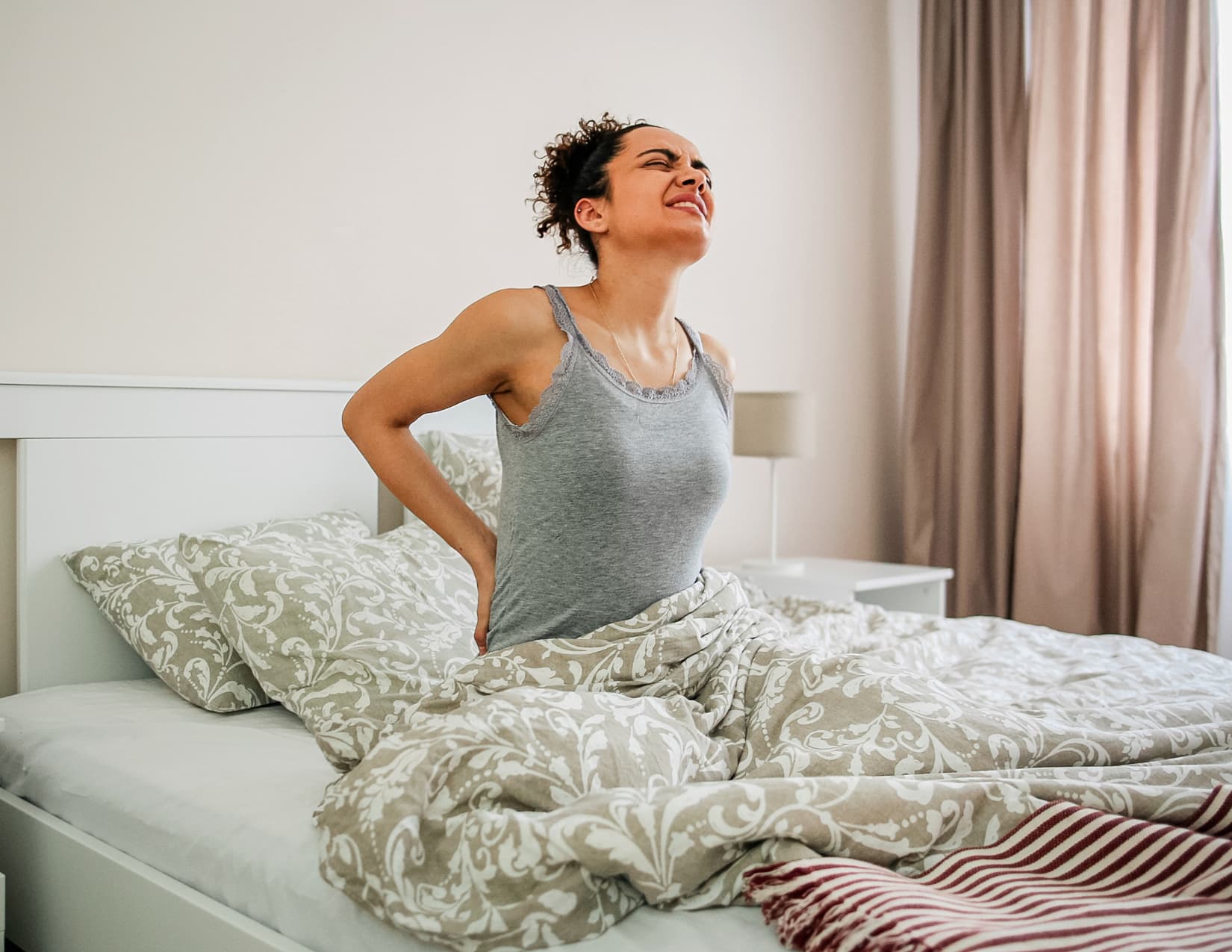 Woman sits up in bed and holds her back, grimacing in pain. 