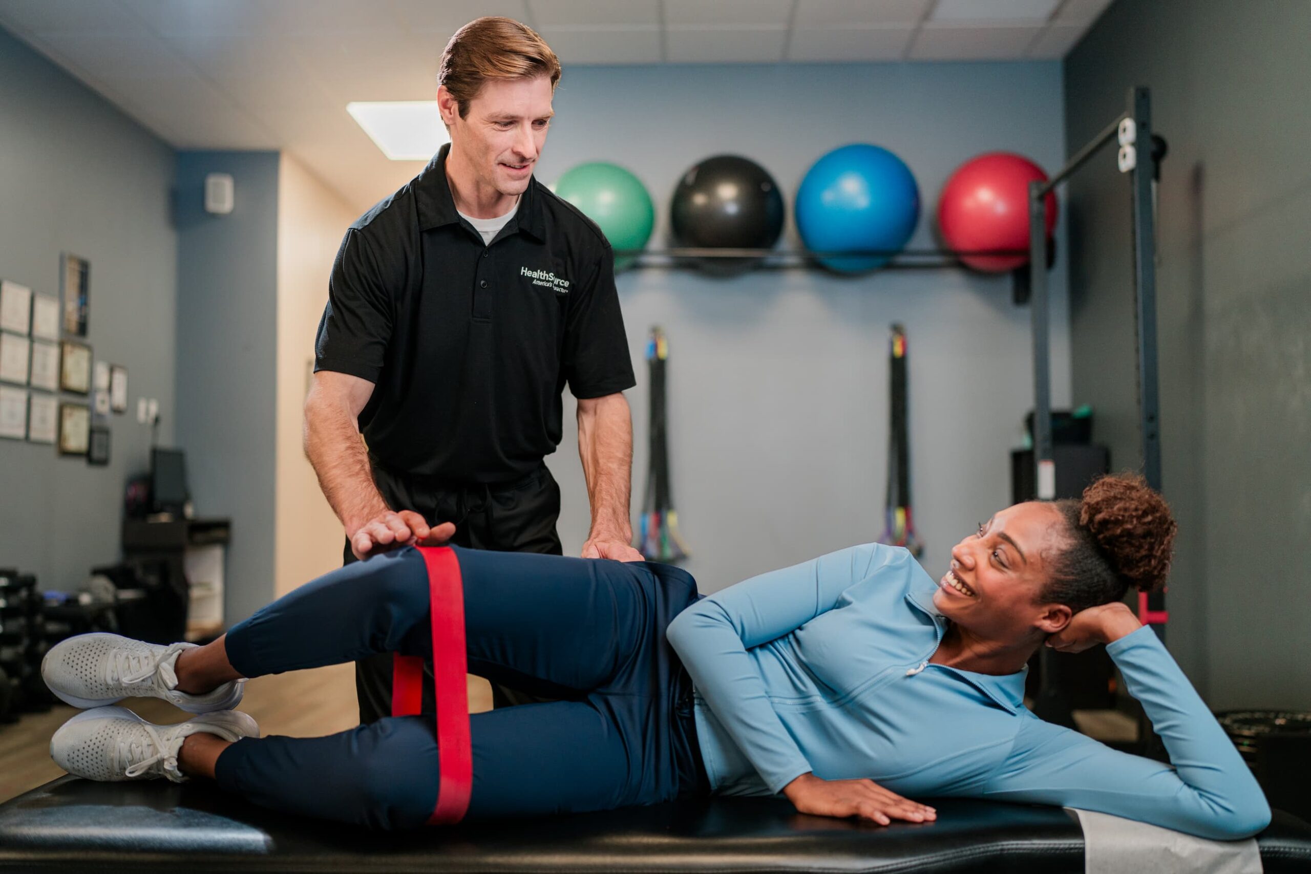 Patient lays on their side, stretching their legs using a resistance band around their thighs. A practitioner guides them. 