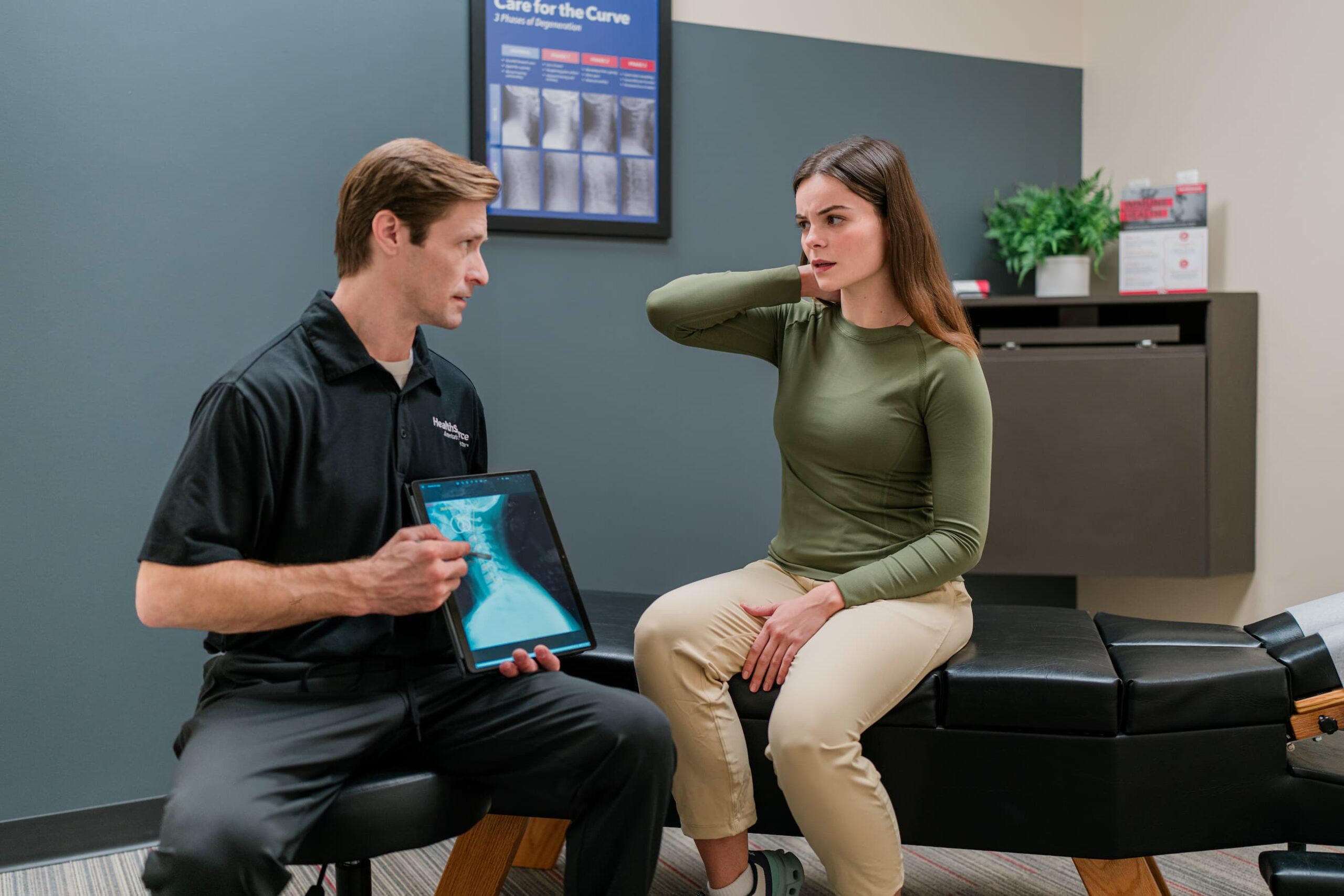 Patient sits at a table, hold their neck and discussing their needs with a practitioner, who holds an x-ray.