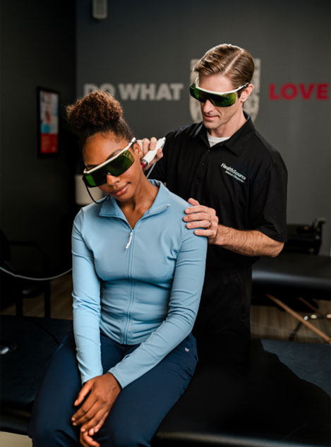 Healthcare professional using a therapeutic device on a seated woman in a clinical setting, both wearing protective green-tinted goggles.