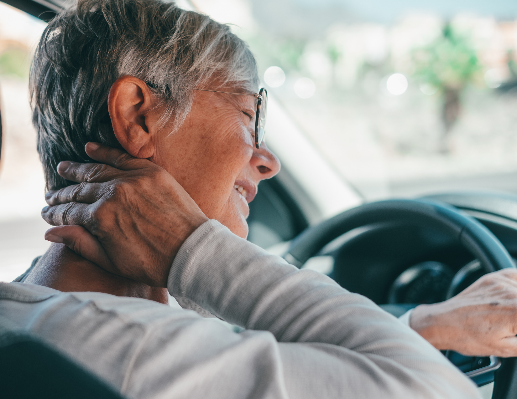 Older woman sits in the driver's seat of a vehicle, holding her neck in pain.