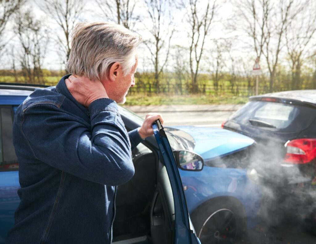 Man holds neck as he stands in the door of his recently crashed vehicle.