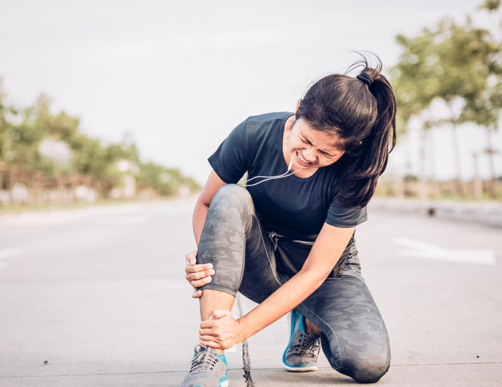 Woman in running gear kneeling on paved sidewalk, clutching twisted ankle