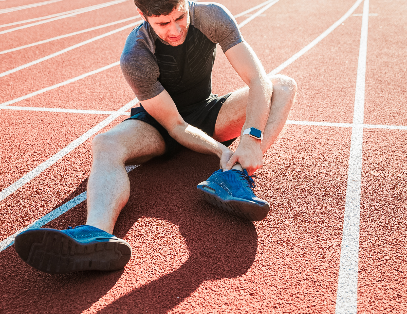 Young man sits on running track, nursing his ankle pain.