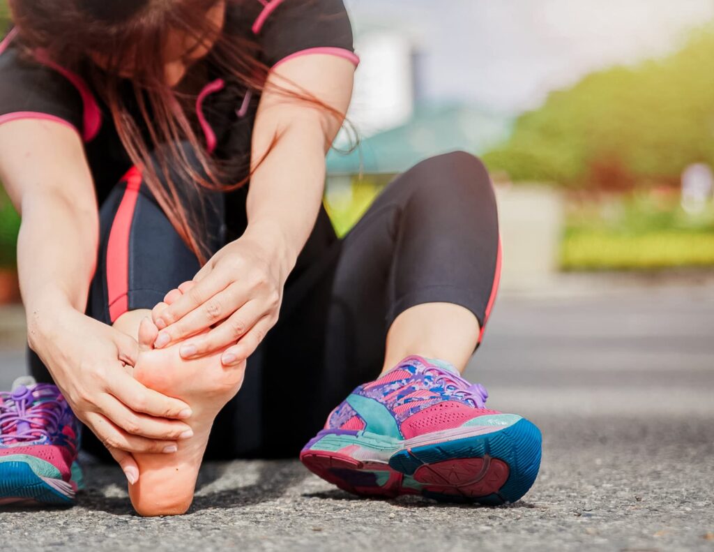 Runner in bright workout gear sits on asphalt and massages her bare foot.