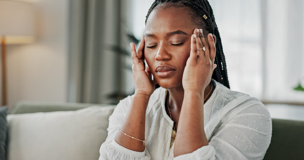 A young woman sitting at home lightly touches her temples, with her eyes closed, as she tries to manage a migraine.