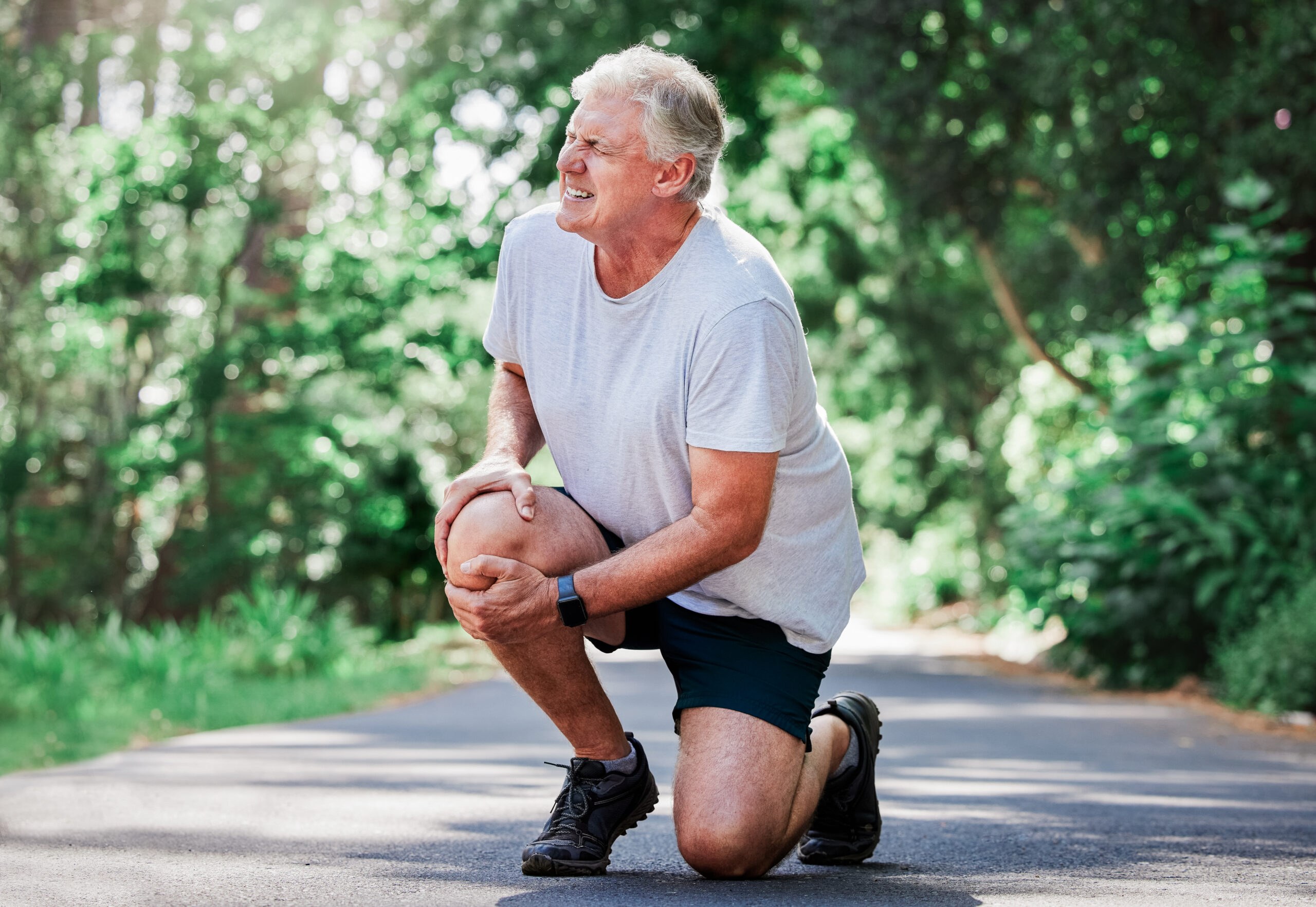 Older man kneels on a running path, gripping his knee and wincing with pain.