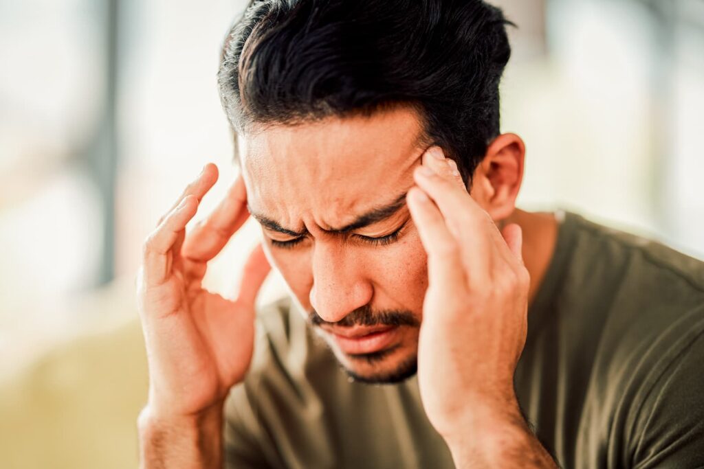 Man with a migraine struggles in a brightly lit room, keeping his eyes closed while lightly pressing at his temples.