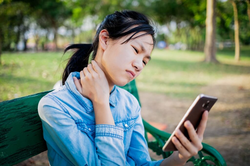 Young woman looks at her phone on a park bench, massaging her neck to assuage pain.