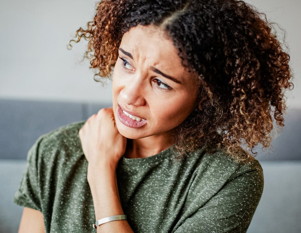 Young woman looks to the left, grimacing in pain while rubbing the area between her neck and shoulder.