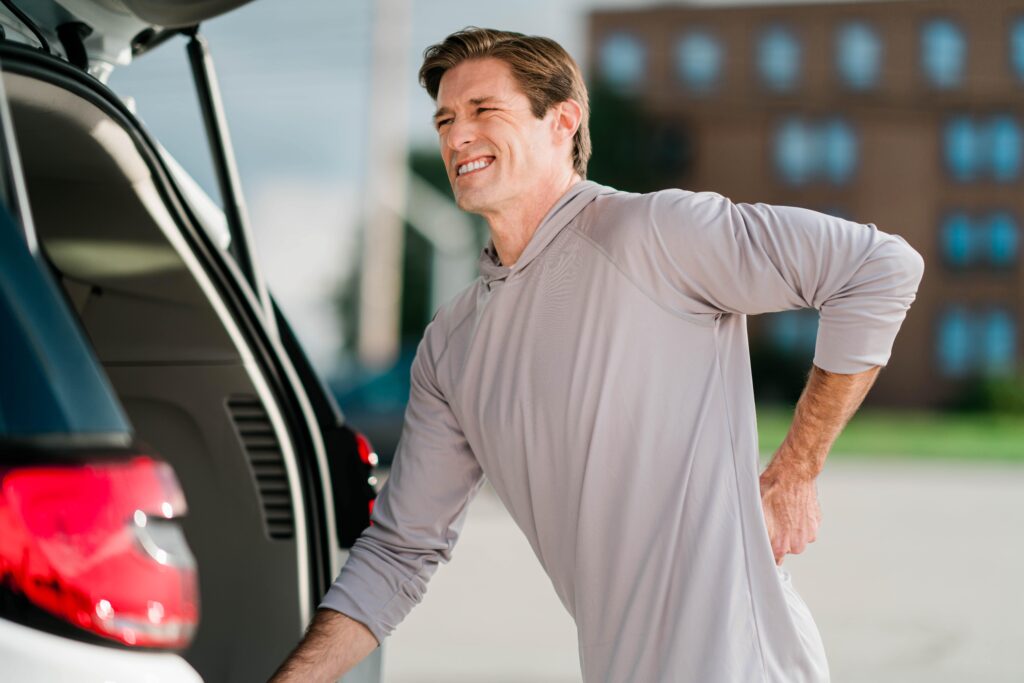 Man winces in pain, holding his back as he leans on the open hatch of an SUV.