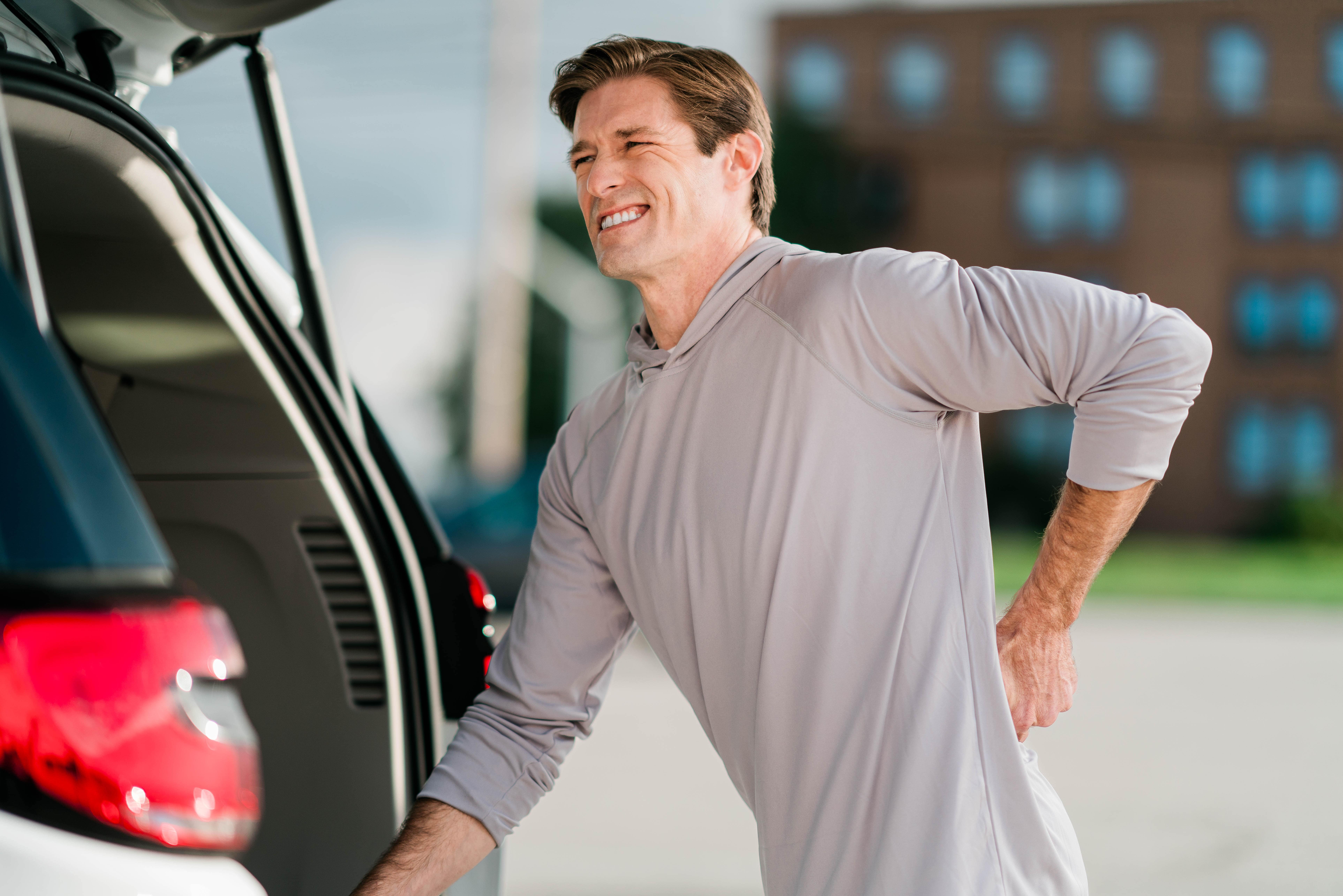 Man winces in pain, holding his back as he leans on the open hatch of an SUV.