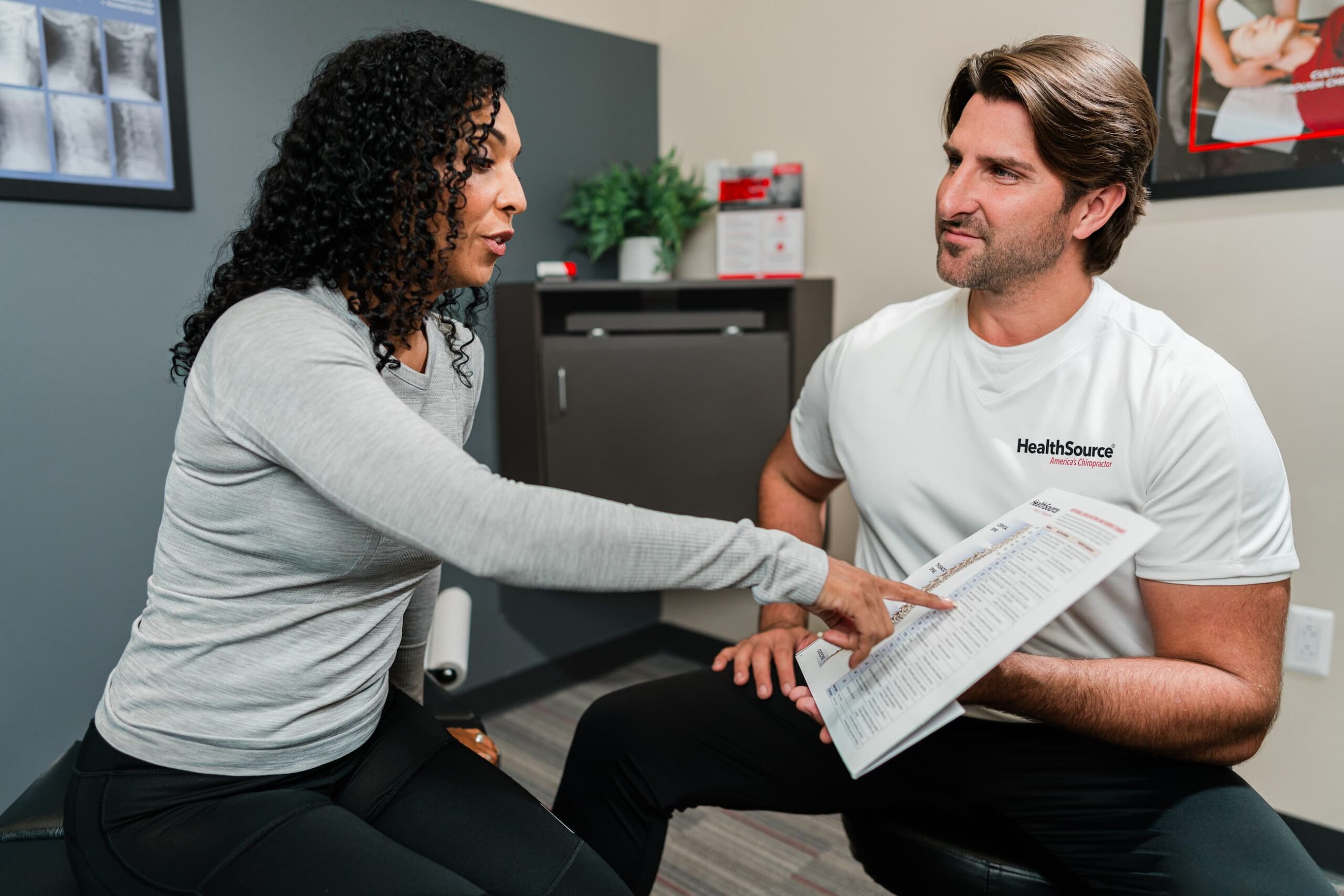 Chiropractor and patient sit down to discuss care. The chiropractor holds a paper up to the patient, who points at the center of the page.