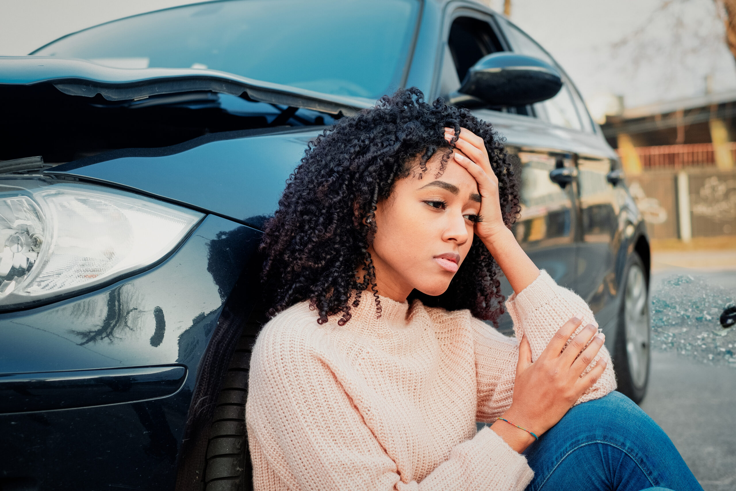 A woman holds her head, sitting next to her car's front tire after an accident.