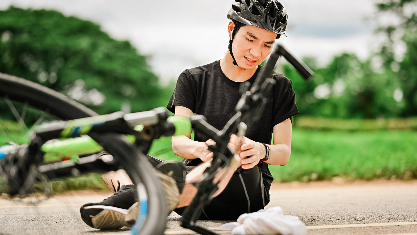 A young man in a black t-shirt and helmet holds his knee after falling off his bicycle.