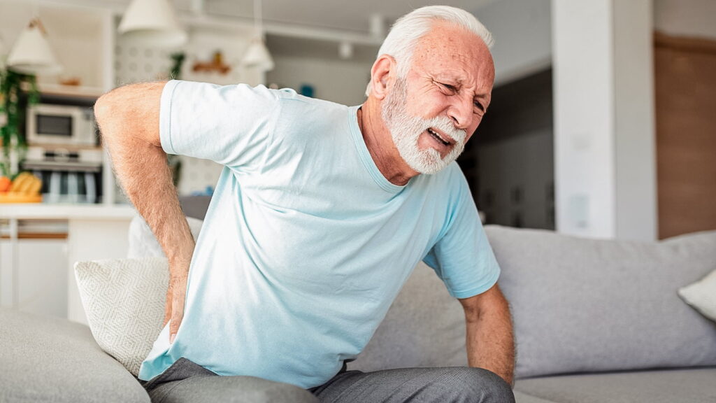 Senior man leans forward on his couch, holding his lower back and wincing due to pain.