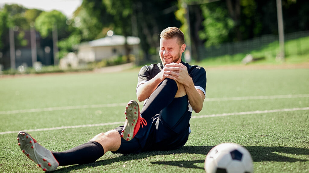 Soccer player sits on the ground, in pain and grimacing, holding his injured knee