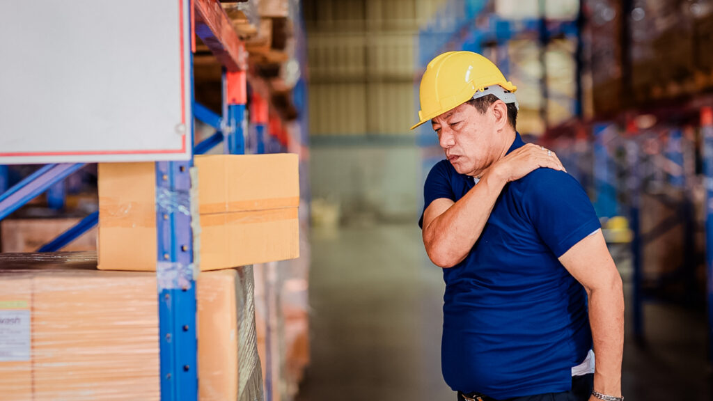 Man in safety gear, standing in an open warehouse, holds his shoulder as he works through old injury pain.