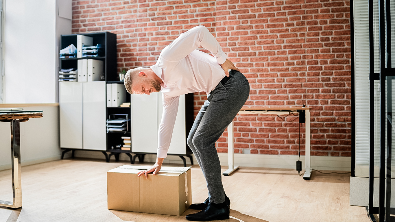 Male employee bends over, leaning on a heavy cardboard box, holding his back and wincing in pain.