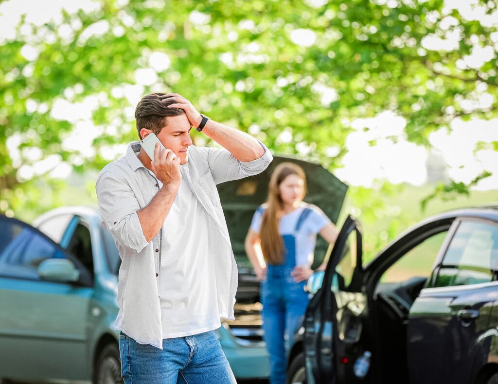 A man stands in front of a car accident, holding his head and talking on the phone, with the other driver standing in the background.