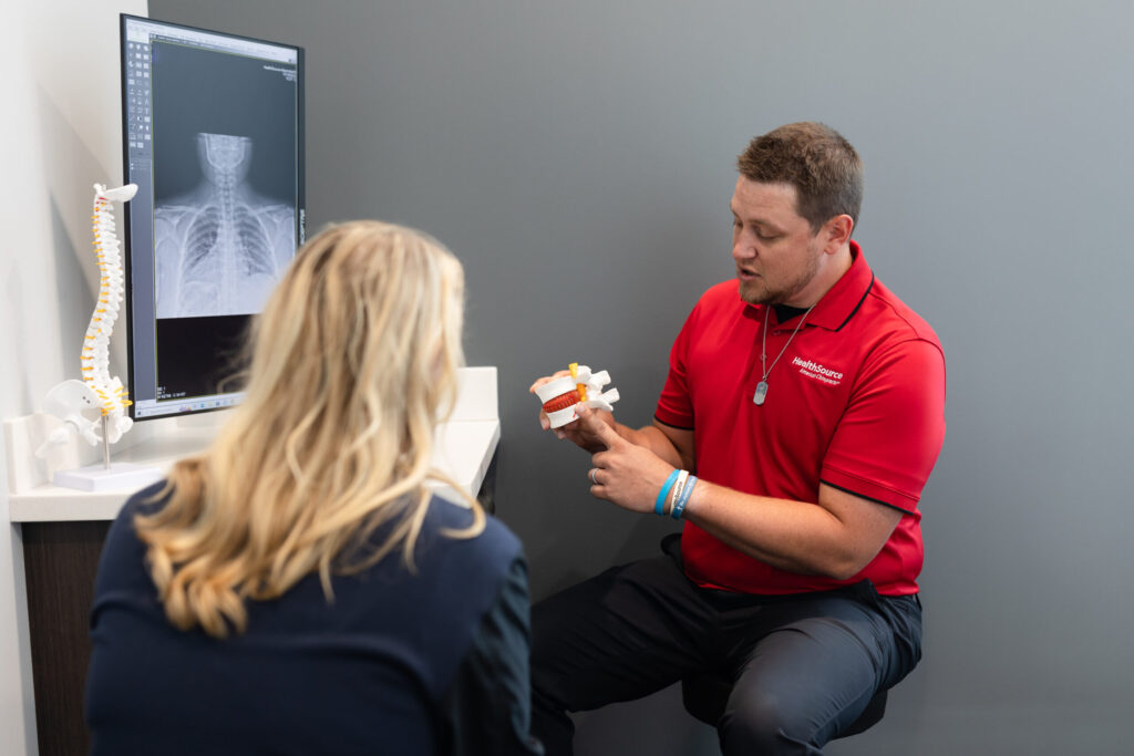 Practitioner sits in a bright office. He points to a model in his hand, identifying the space between vertebrae for a patient.