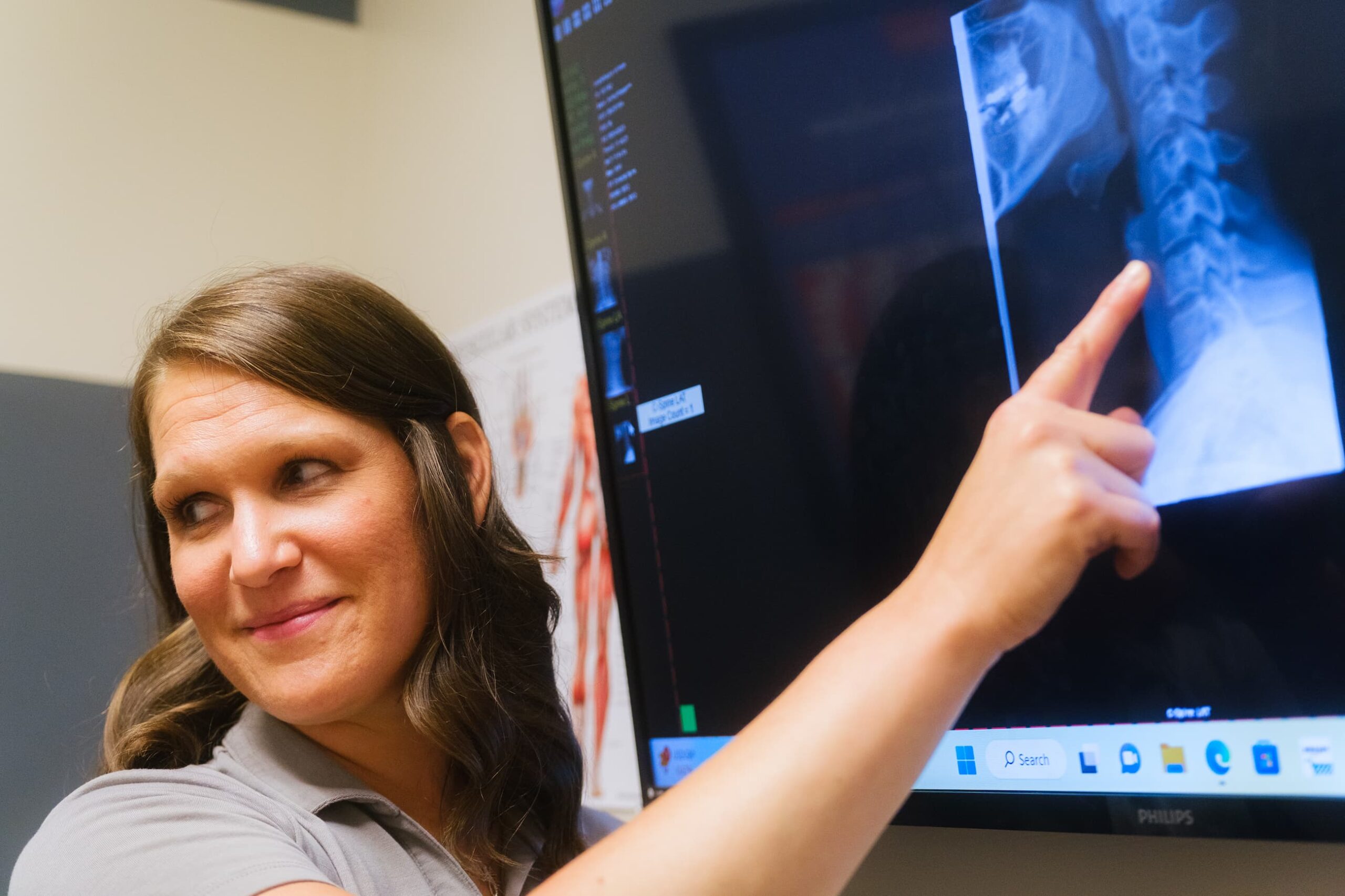 Practitioner stands near a screen, pointing at an x-ray of a patient's neck.