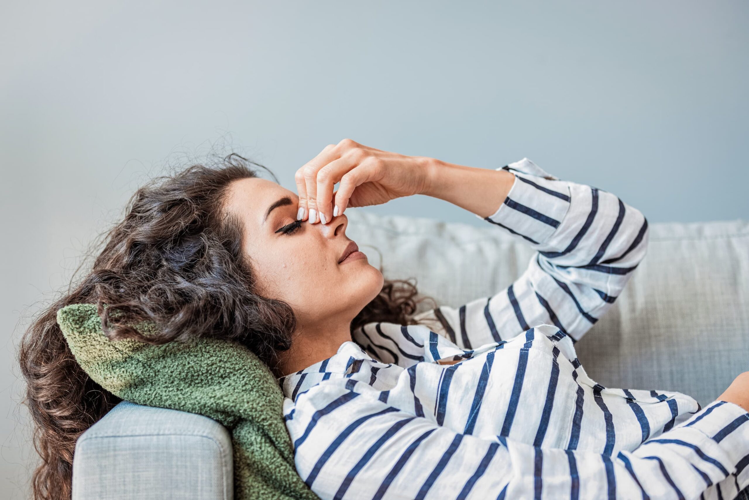 Young woman lays back on couch, rubbing the bridge of her nose as she experiences migraine pain.