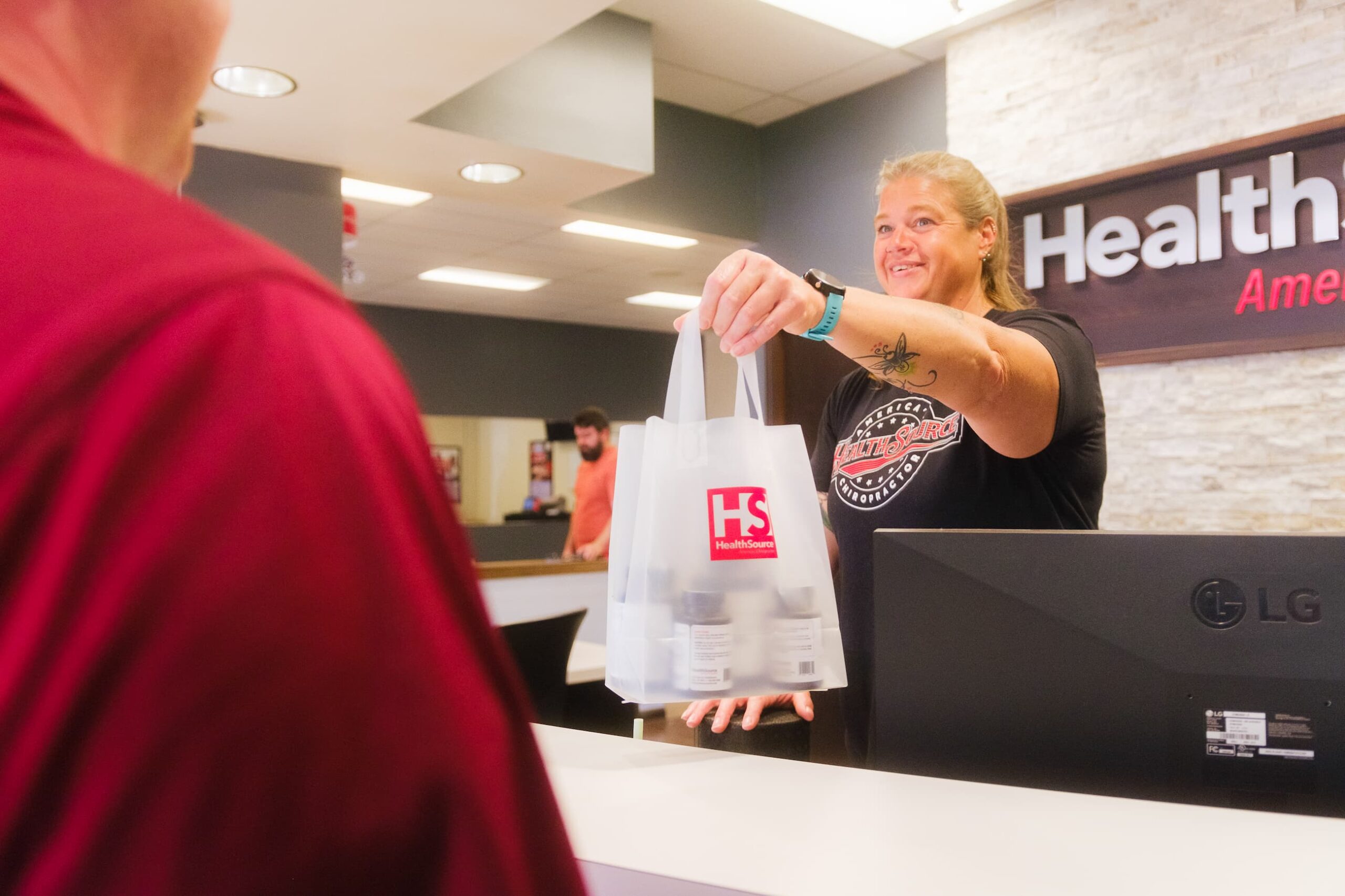 HealthSource employee standing behind a cash register passes a branded HS bag full of supplements to a patient.