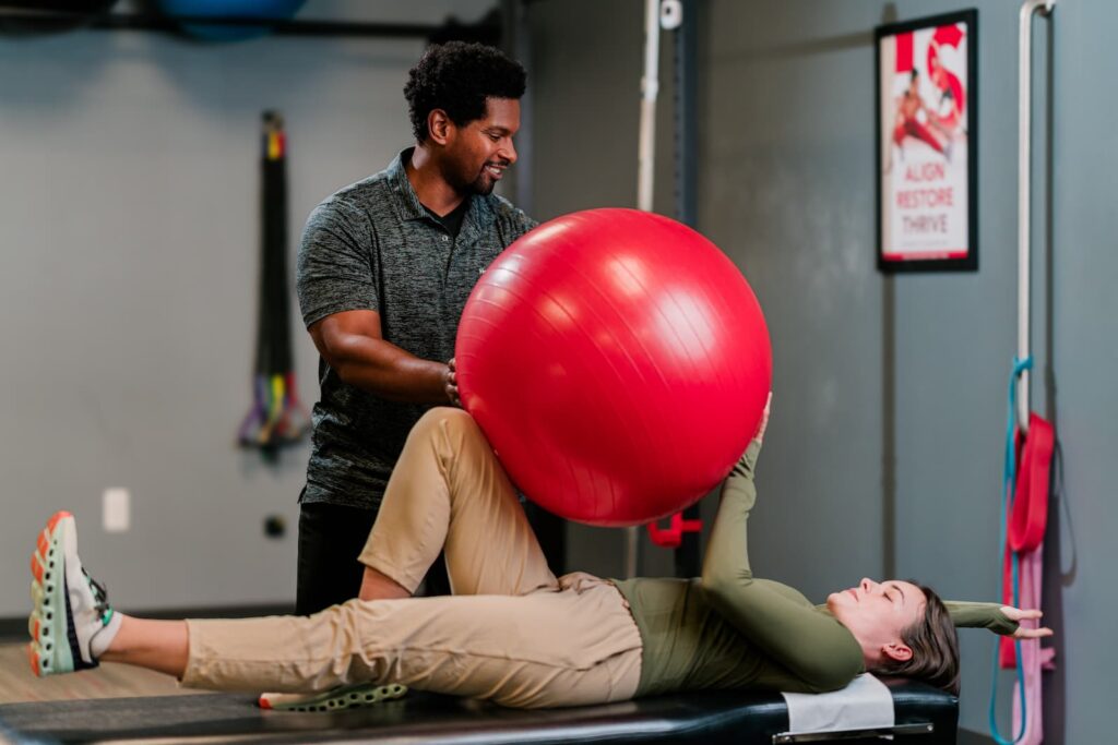 Patient lays on exam table, holding a large red ball between her hands and right knee while a practitioner assists.