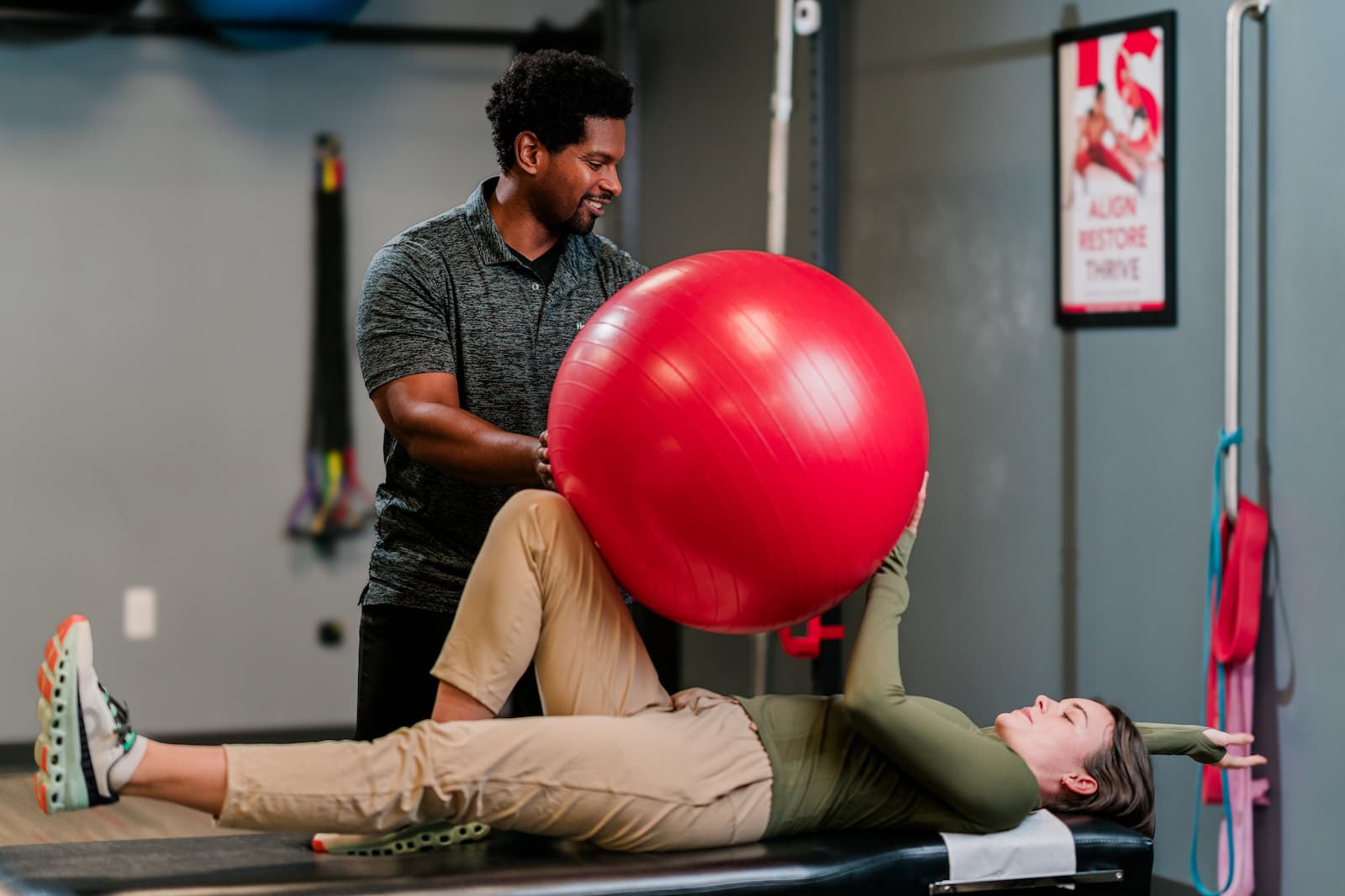 Patient lays on exam table, holding a large red ball between her hands and right knee while a practitioner assists.