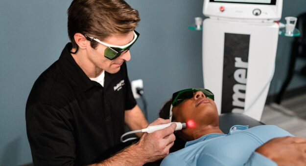 Practitioner applies laser treatment to a patient's jawline, near their ear. Both practitioner and patient wear protective eye gear.