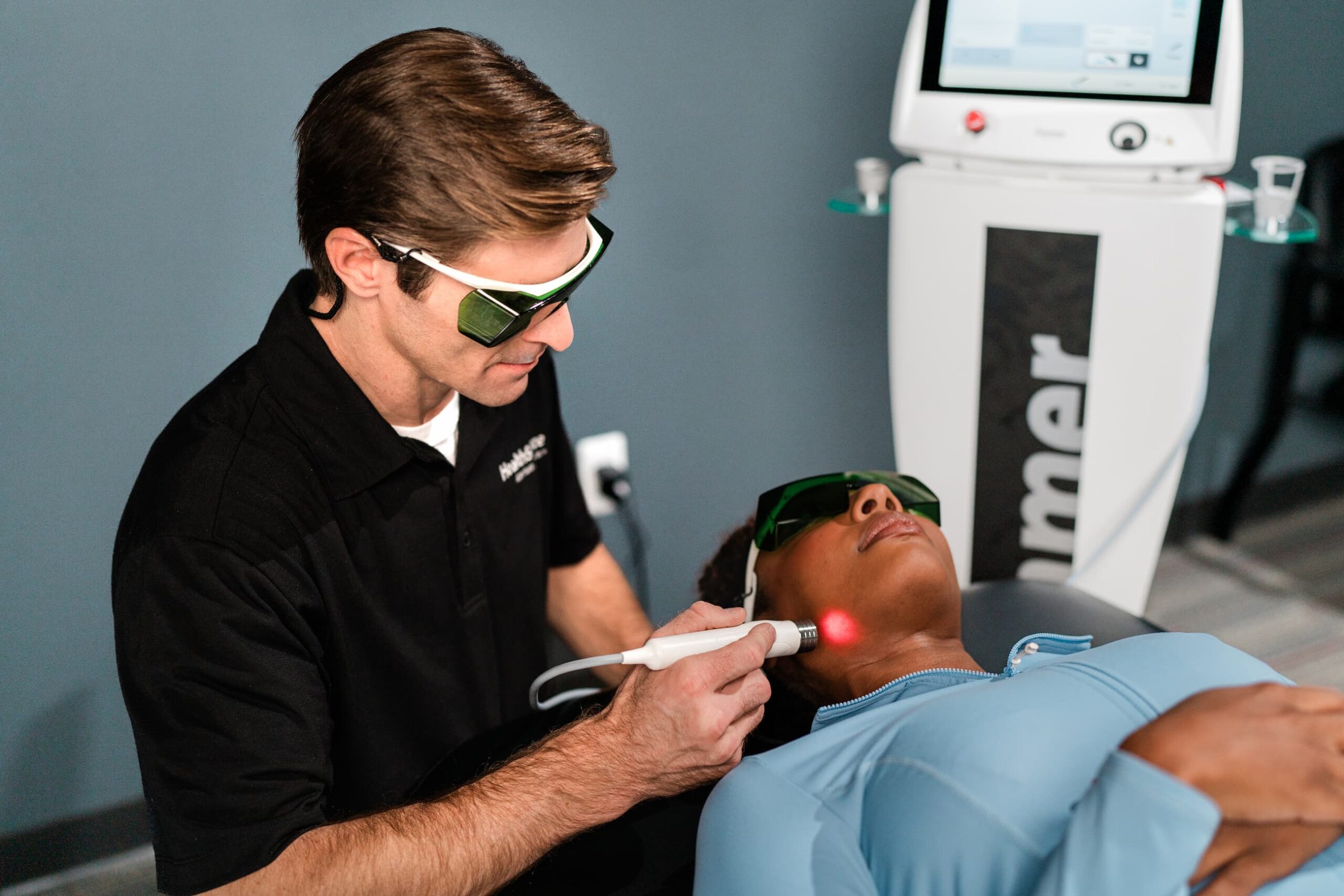 Practitioner applies laser treatment to a patient's jawline, near their ear. Both practitioner and patient wear protective eye gear.