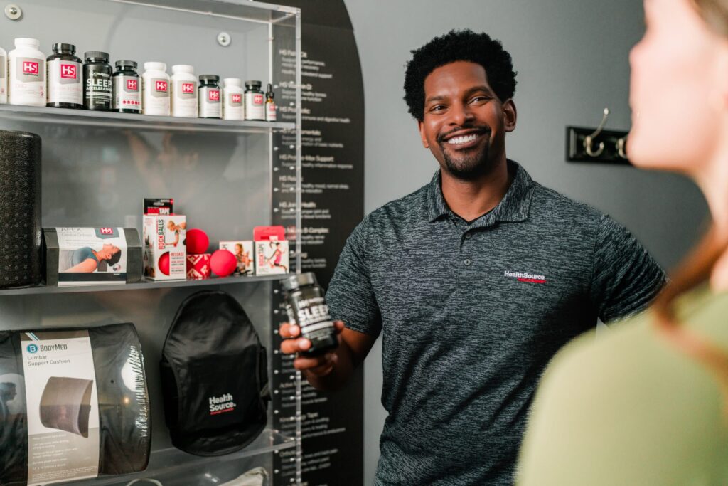 HealthSource practitioner shows a patient a bottle of supplements, pulled from a full shelf displaying an array of other supplements.