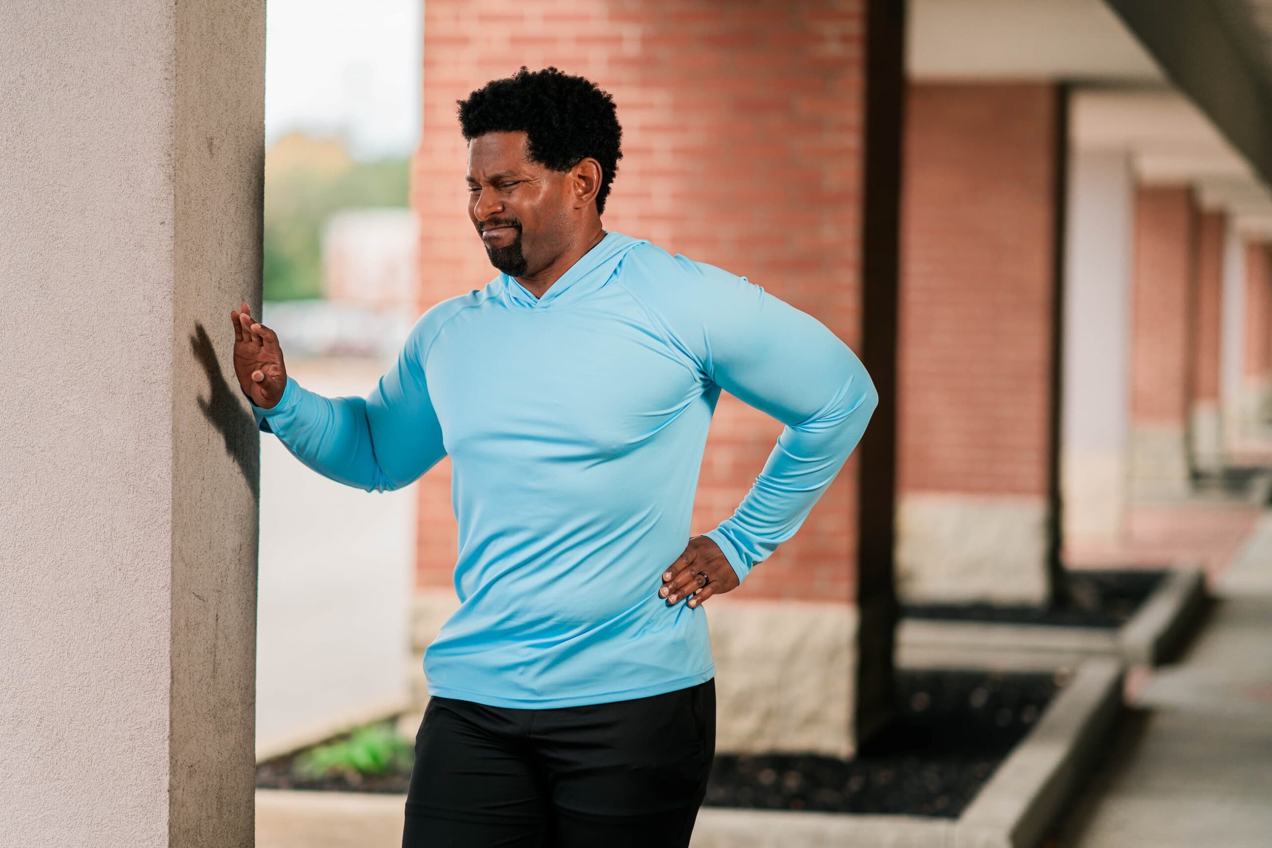 Man in athletic wear leans against a column of a building complex, gripping his left hip in pain.