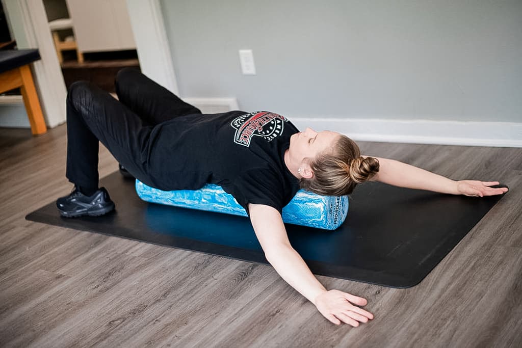 Patient lays on a foam roller, stretching their arms above their head and resting them on the ground.