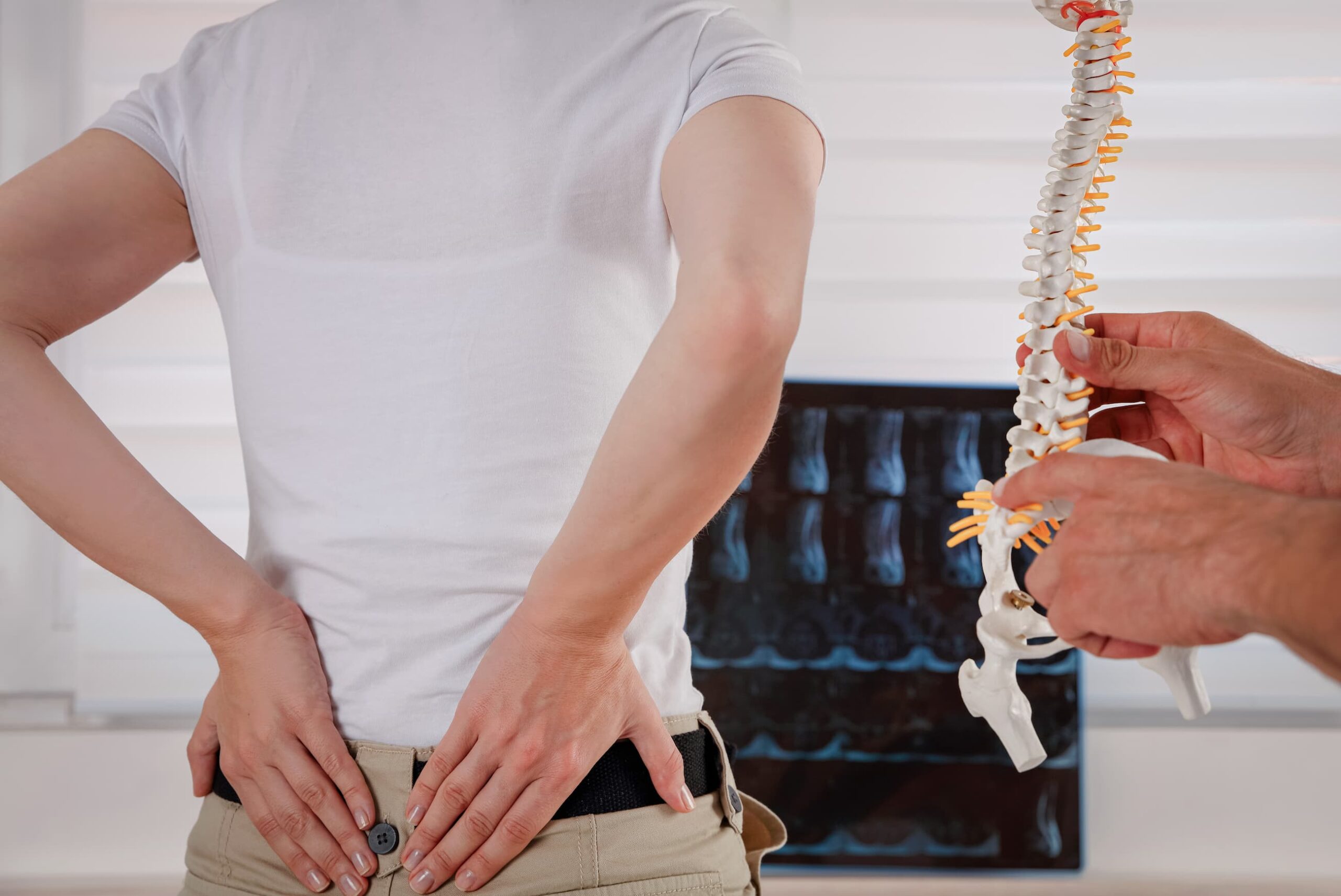 Woman in a medical office presses her hands to her lower back while a doctor points to the same area on a skeleton model.