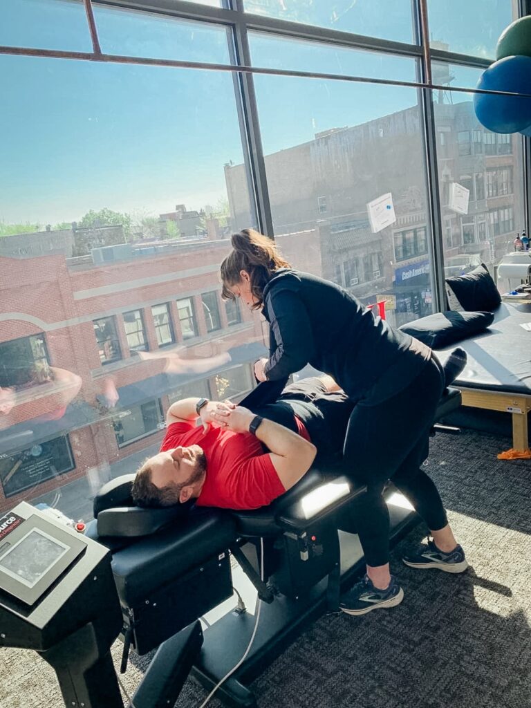 A practitioner assists a patient on a spinal decompression, set beside large windows letting in bright sunlight.