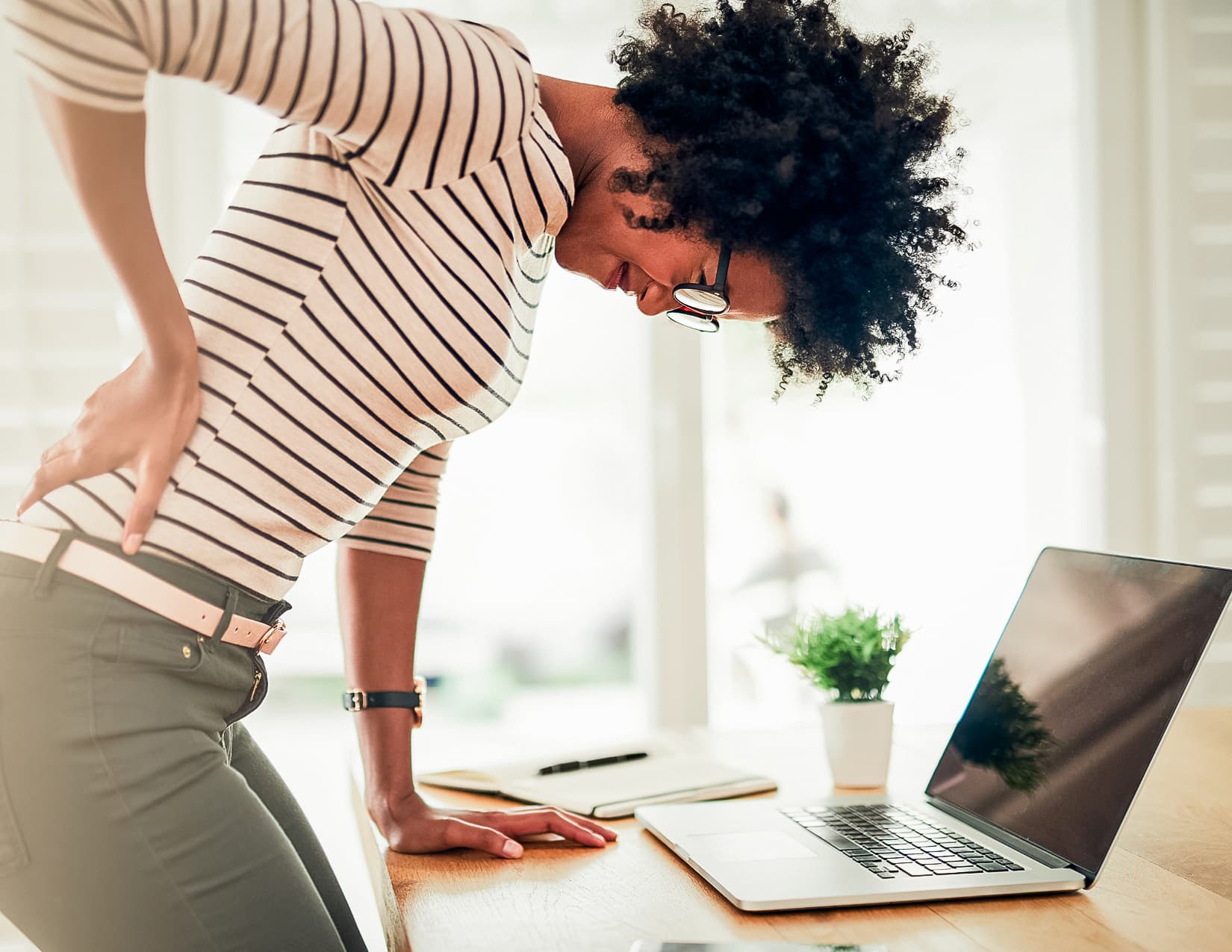 Young office worker leans on their desk, gripping their lower back and grimacing. 