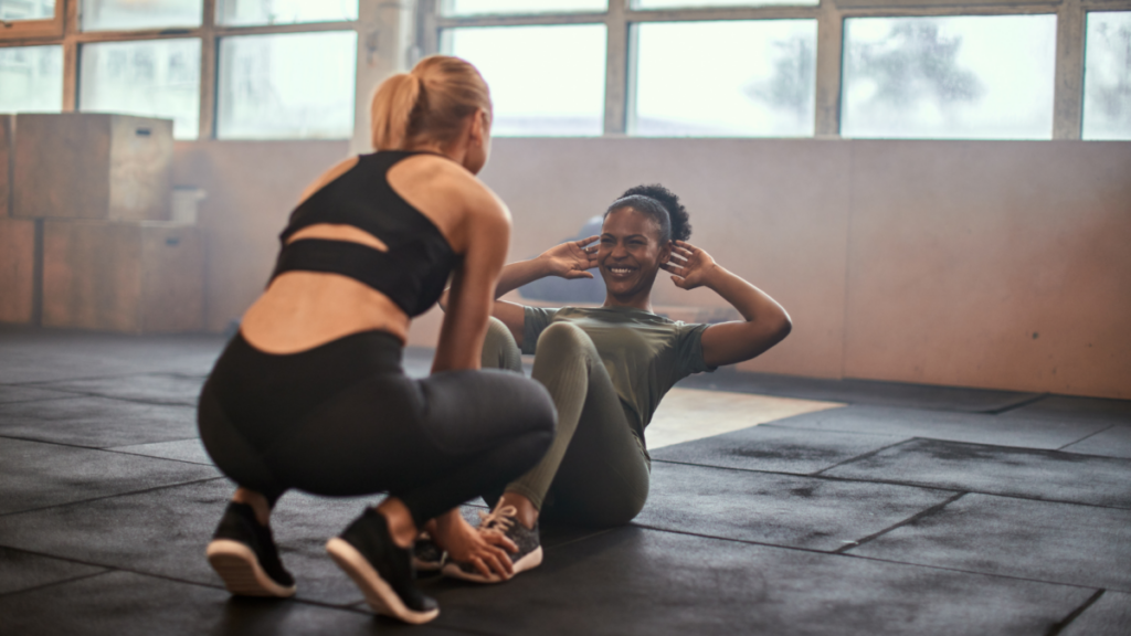 Two women exercising in a gym. One woman is performing sit-ups on a workout mat while smiling, and the other is crouched in front of her, holding her feet for support.