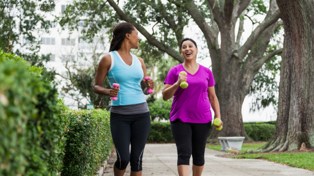 Two women walking outdoors on a tree-lined path, holding small hand weights and smiling as they engage in conversation while getting their 10,000 steps a day. 