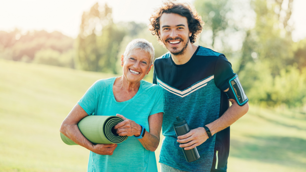 Two people standing side by side in a park, smiling warmly. One person is holding a rolled-up yoga mat, while the other holds a water bottle, suggesting they are ready for outdoor exercise or yoga.