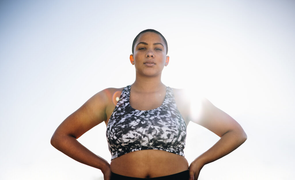 A person in sportswear standing outdoors with their hands on their hips, feeling confident from the benefits of chiropractic care, with the sun shining from behind.