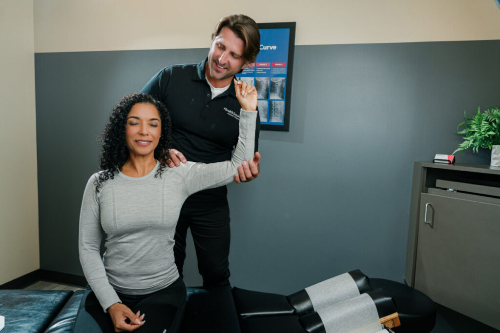 A patient sits on an examining table as a HealthSource chiropractor examines her shoulder