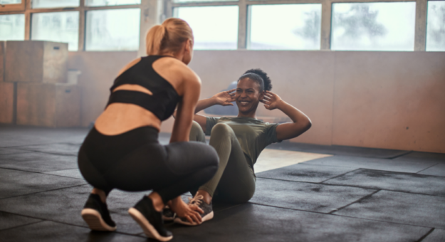 Two women exercising in a gym. One woman is performing sit-ups on a workout mat while smiling, and the other is crouched nearby, holding her feet for support.