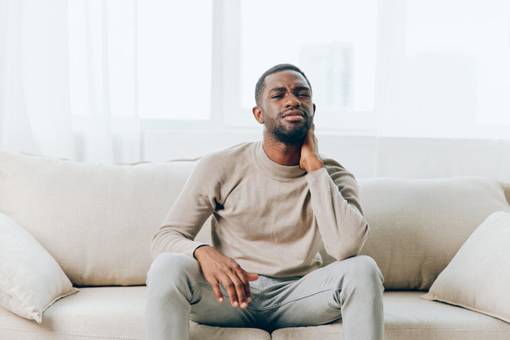 Stressed man with a headache sitting alone on the couch at home, holding the side of his head and neck. He has a clear expression of pain and frustration on his face.