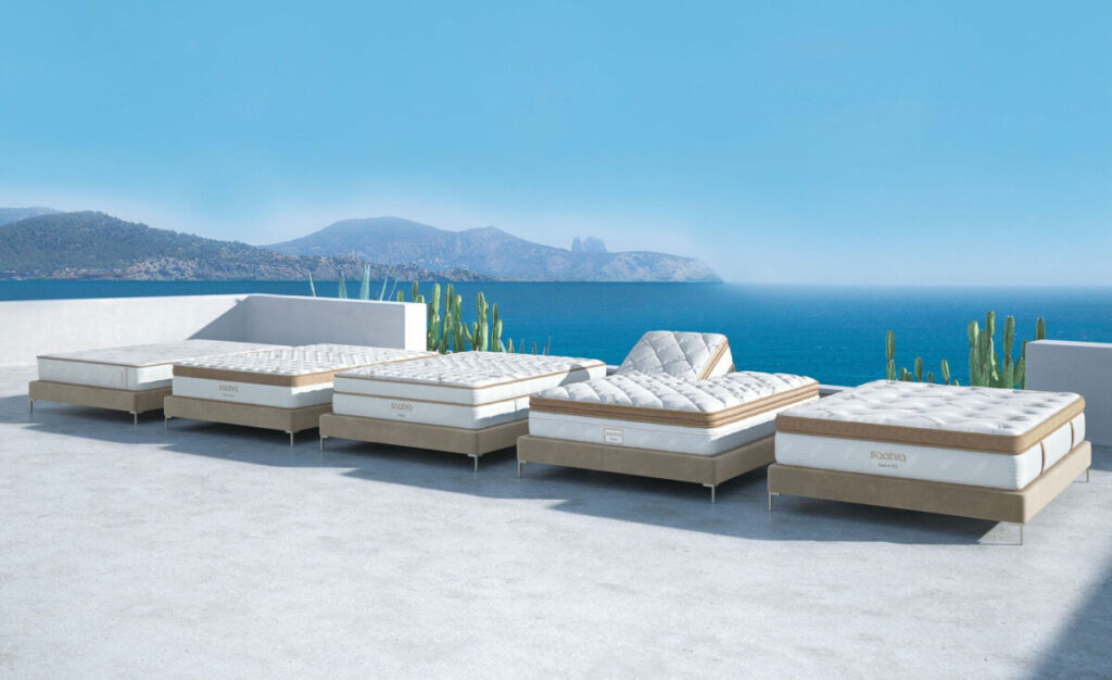 Five Saatva mattresses without bedding, placed on a platform overlooking a blue ocean, mountains, and a clear blue sky. One mattress is in an upright position, showcasing its adjustable top feature.