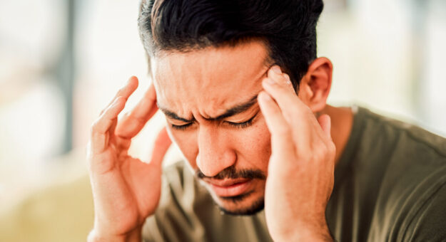 A man with dark hair and a beard is experiencing a headache, pressing his fingers against his temples with a pained expression. His eyes are closed, and his brow is furrowed. The background is blurred, suggesting an indoor setting with natural light.