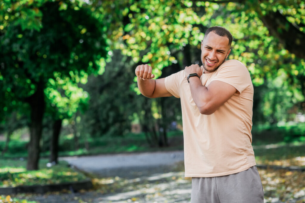 A person in a park, standing and holding their right shoulder with their left hand while raising their right arm, showing a grimace of pain and discomfort.