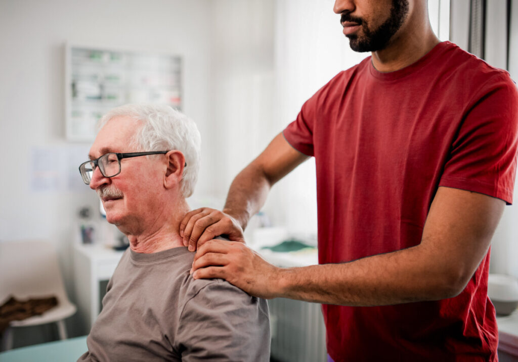 An older person sitting relaxed in front of a chiropractor, who is using both hands to perform an adjustment on the person's left shoulder.