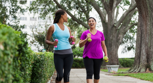 Two women walking outdoors on a tree-lined path, holding small hand weights and smiling as they engage in conversation.